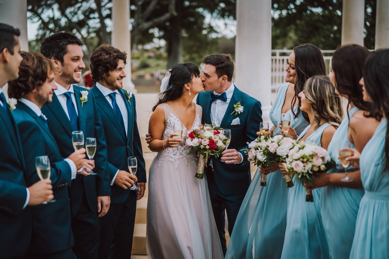 A wedding couple is kissing surrounded by bridesmaids and groomsmen